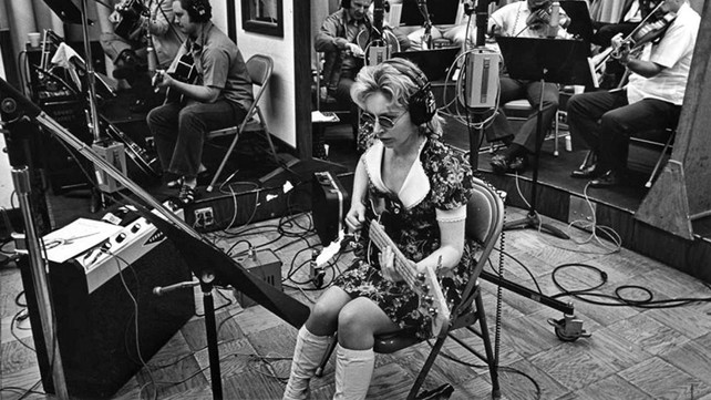 Carol Kaye in a session. She is holding the bass, preparing to play. She is the only woman in the room. Image is in black and white. 